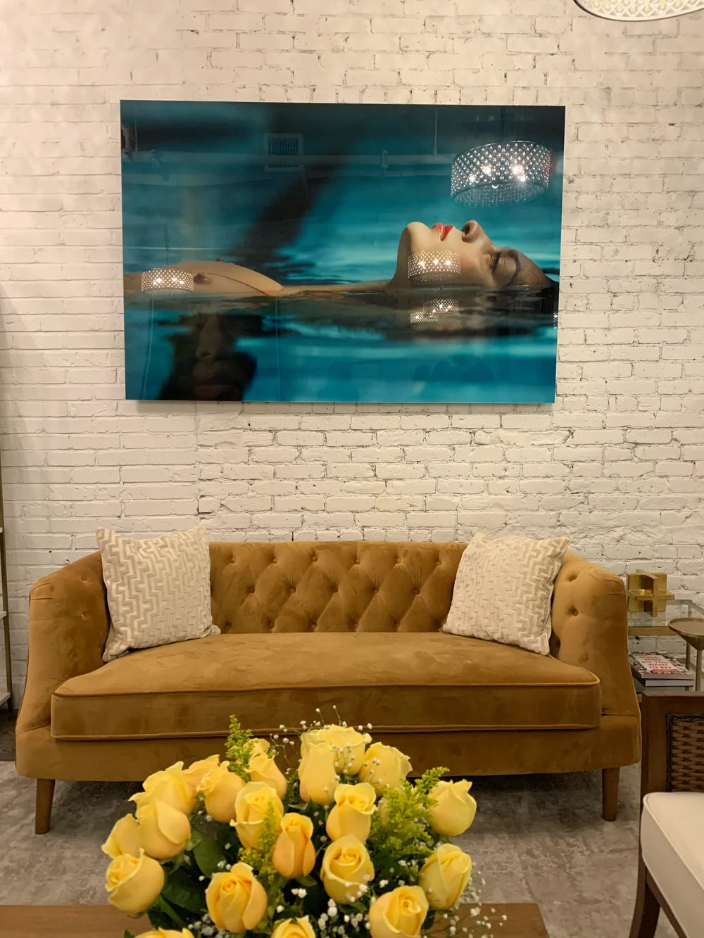Face-mounted photograph of a woman floating in turquoise water, hung on exposed brick above a velvet sofa